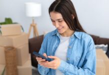 A young woman wearing a denim shirt looks at her phone with a stack of cardboard boxes and a couch behind her.