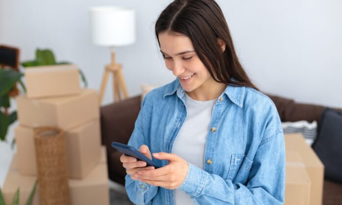 A young woman wearing a denim shirt looks at her phone with a stack of cardboard boxes and a couch behind her.