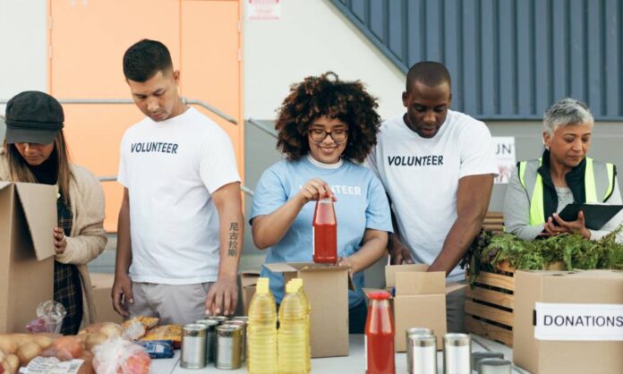 A group of young volunteers are placing groceries into food boxes. Their supervisor is holding a clipboard and taking notes.