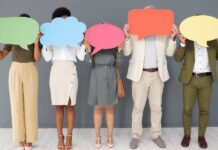 A diverse group of five people in business attire standing against a gray wall and holding speech bubble cut outs.