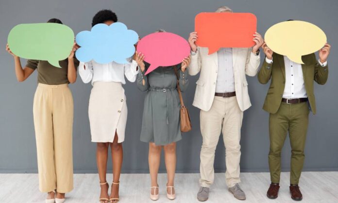 A diverse group of five people in business attire standing against a gray wall and holding speech bubble cut outs.