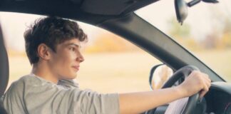 A young man wearing a sweatshirt driving a sedan with one hand on the wheel and his other arm bent, leaning on the car door.