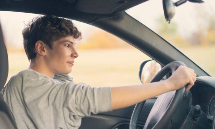 A young man wearing a sweatshirt driving a sedan with one hand on the wheel and his other arm bent, leaning on the car door.