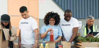 A group of young volunteers are placing groceries into food boxes. Their supervisor is holding a clipboard and taking notes.