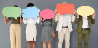 A diverse group of five people in business attire standing against a gray wall and holding speech bubble cut outs.