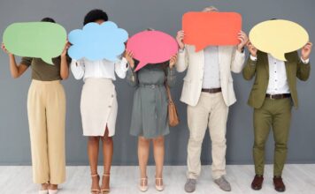 A diverse group of five people in business attire standing against a gray wall and holding speech bubble cut outs.