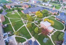 An aerial view of a college campus with red brick buildings, sidewalks, a parking lot, trees, and green grass.