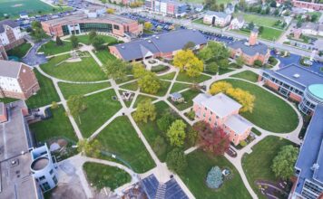 An aerial view of a college campus with red brick buildings, sidewalks, a parking lot, trees, and green grass.