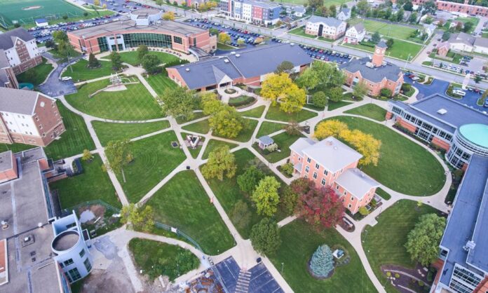 An aerial view of a college campus with red brick buildings, sidewalks, a parking lot, trees, and green grass.