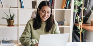 A young woman sitting at a desk, surrounded by papers and a coffee cup, smiling as she types on her laptop.