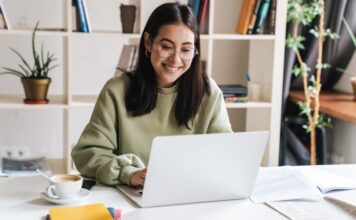 A young woman sitting at a desk, surrounded by papers and a coffee cup, smiling as she types on her laptop.