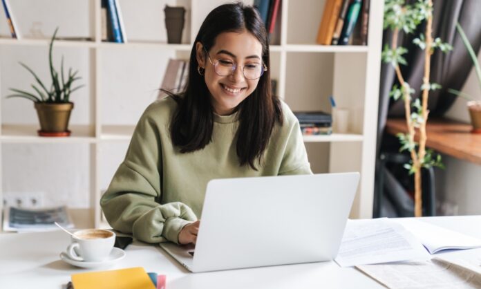 A young woman sitting at a desk, surrounded by papers and a coffee cup, smiling as she types on her laptop.