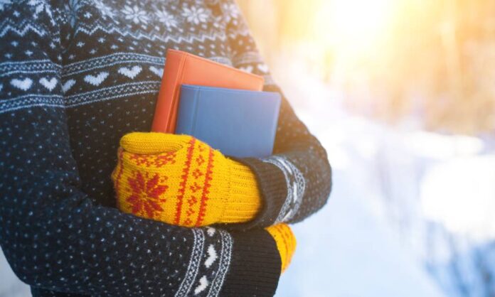 A person in a clack and white sweater and yellow and red knit mittens holds two books outside.